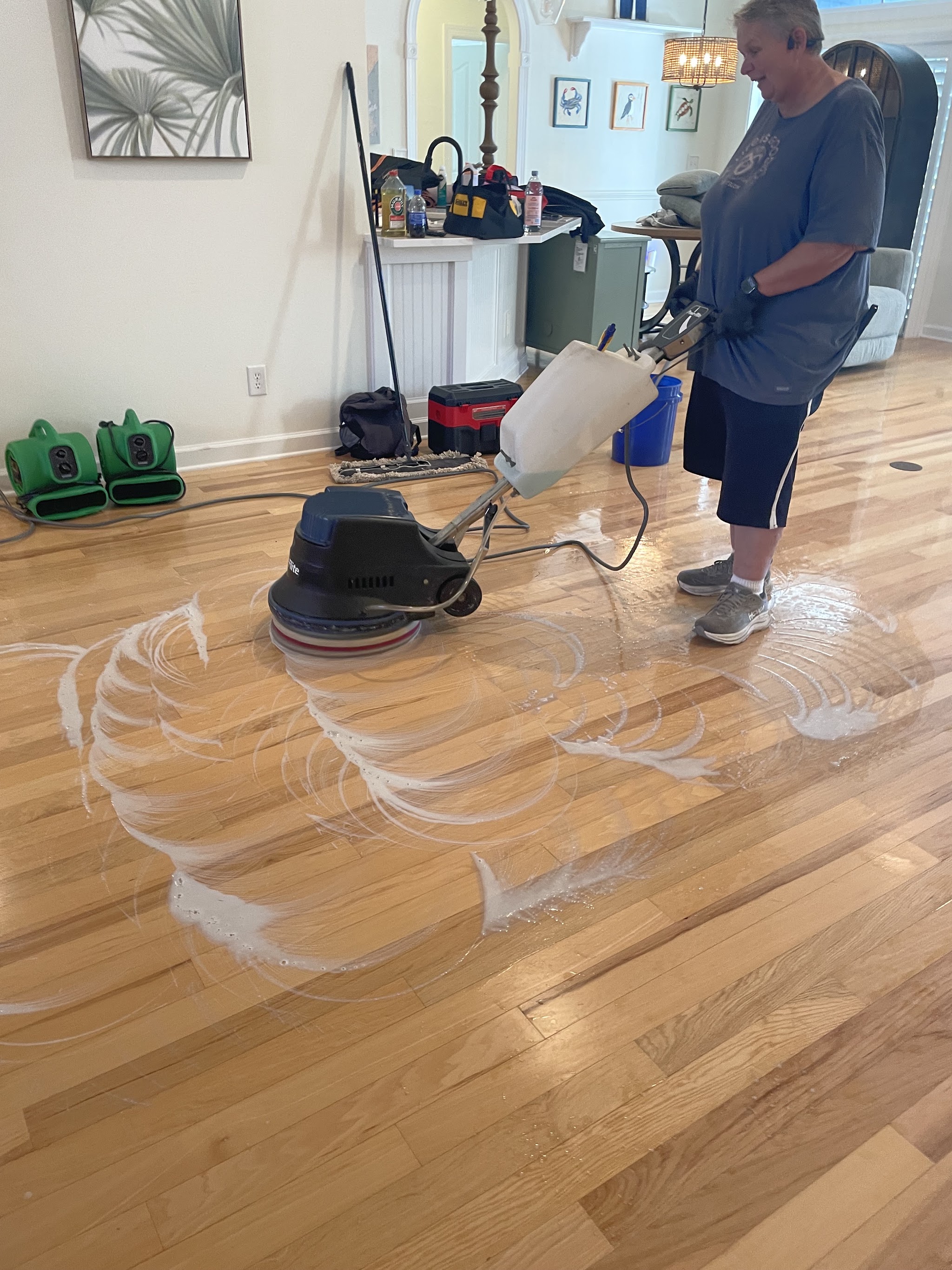 A cleaner working a floor machine in a bright home, with drying fans and equipment set up behind